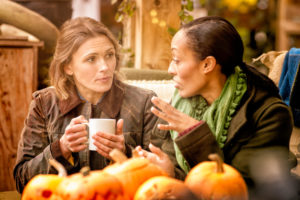 Two friends sit in cafe in front of display of pumpkins and drink coffee and have conversation