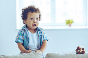 Child's face crumples with mouth open as he looks off to the side of photo from back of sofa
