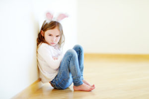 Very angry child with long hair and rabbit ear costume sits on floor with arms crossed, glaring