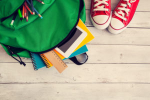 Green backpack lies open, school supplies sticking out of the top, on wood floor next to red sneakers