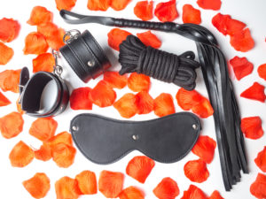 Photo of black leather handcuffs, blindfold, flogger, and rope on flower petal background
