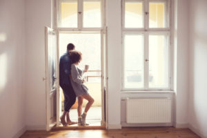 Couple leans into each other in front of open door by walls of windows in bright house