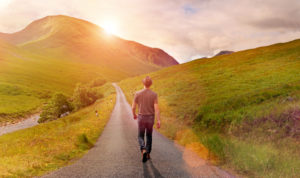 Rear view of person in casual clothes and hat walking alone on a narrow country road through a green valley surrounded by mountains, heading toward the rising sun. Warm sunlight with lens flare. Taking decisions and choosing a direction for new beginnings, following the way forward, not looking back.