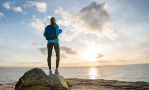 Hiker with backpack and long ponytail watching the sunset on top of a mountain and over the ocean