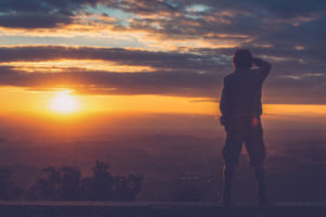 Rear view photo of person with short hair wearing jacket and shorts standing on ledge looking out at sunset 