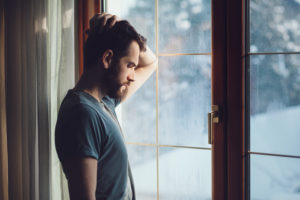 Young adult with short hair, facial stubble, leaning on one arm looking out windowed door during rain