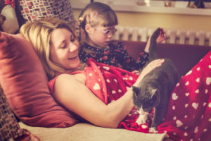 Parent and young child with glasses, barrettes, braids sit on daybed and pet cat