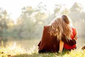 Rear view of two young adults with long hair sitting on grass. one hugs the other