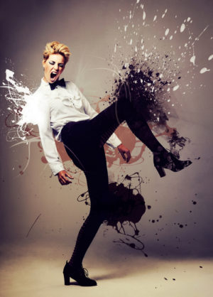 Tall person with short hair wearing bowtie, shirt, slacks, and heeled boots kicks out against splashes of paint