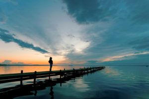 Young person with long hair standing on lakeside on jetty having a look at magical cloudscape and sunset colors.