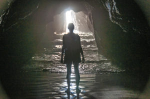 Person stands in shallow water looking up stone steps water is coming over looking up at light from tunnel exit