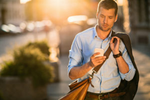 Person walking down sunny street looks at phone, holding coat over shoulder with one hand