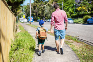 Rear view of parent walking child with backpack and hat down tree-lined street
