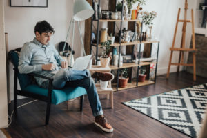 Young adult with short hair wearing jeans and button-down sits in chair and works on laptop