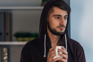 Portrait of pensive young adult in hooded shirt holding coffee cup and looking away while standing at home