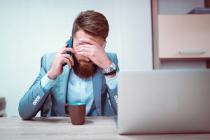 Person sitting at desk in small office with beard and short hair covers face with hand while holding phone to ear