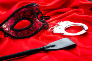 Riding crop, handcuffs, and lace mask lie on red satin