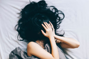 Person with long black hair lies in bed, hands and hair over face