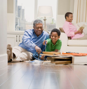 Child helps grandfather build furniture while child's parent sits reading on sofa in background