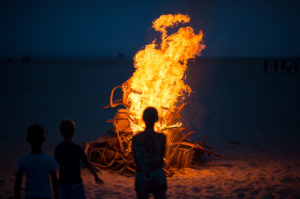 Group of children around campfire at beach, one child in center staring at large fire but from a distance
