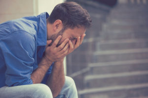 Person with short hair in work shirt crying on stone steps outside, covering face in hands