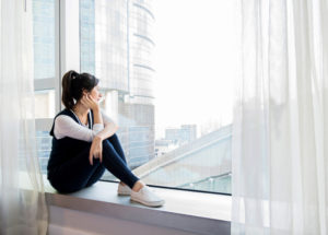 Young adult with ponytail in casual comfortable clothes sits on windowsill, leaning cheek on hand and looking out