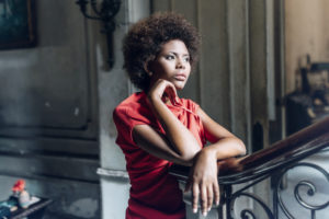 Young adult in red blouse with natural hair stands leaning on banister, lost in thought, with serious expression