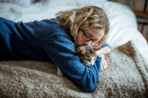 Person in blue sweater with long wavy blonde hair and glasses lies on bed with somber expression holding cat and cuddling 