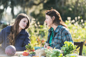 Mother and daughter sitting at a table outdoors on an organic farm, conversing on a bright, sunny day. On the table is fresh produce, including purple cabbage, radishes, leafy green vegetables and a basket of eggs.