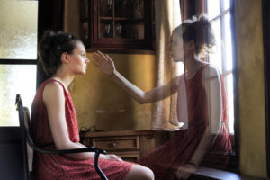 Young adult in red dress with hair pulled back sits somberly in chair with ghostly twin reaching out toward her