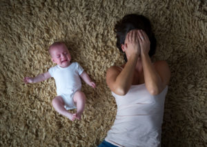 Stressed mother lies on back on carpet next to fussing baby, covering face with hands in distress