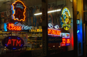 Photo shows neon signs advertising various alcoholic beverages in window of street storefront at night