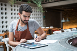Young person with dark hair and business apron works at bar table on tablet 