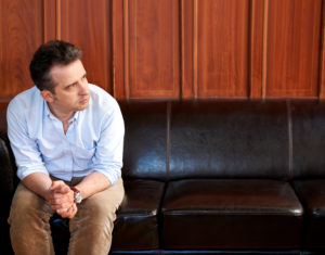 Shot of person of middle years with short hair in business dress sitting on sofa, hands folded, looking to one side