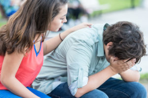 Young teen leans forward with face in hands while friend places comforting hand on back