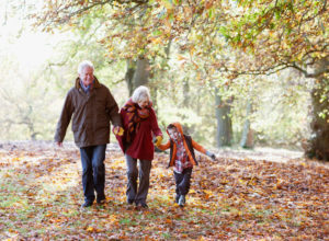 Mature couple walks along path strewn with autumn leaves holding hand of young grandchild