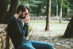 Adult with short dark hair and beard sits on bench, hands steepled against face, looking down