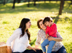 Grandparent, parent with long hair and white blouse, and young child sit together laughing in park at picnic