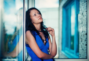 Person in blue lacy top with long dark hair leans back against wall, holding necklace, thinking