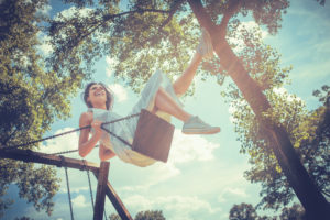 Young person with long hair in dress and sneakers laughs and swings feet in tree swing