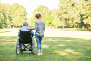 Rear view photo of caregiver with hair in bun standing next to person in wheelchair, hand on their shoulder