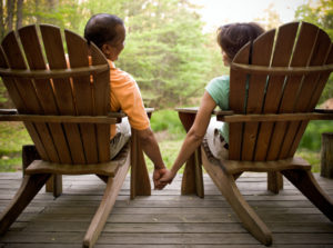 Rear view photo of older couple sitting on deck in Airondack chairs holding hands