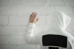 Photo shows back of young teen wearing a hoodie standing with palm on whitewashed wall