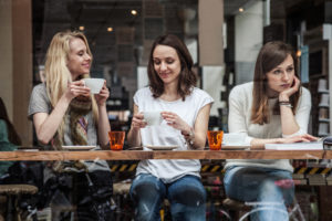 Group of three sit at cafe table. Two talk and laugh while one looks down and away.