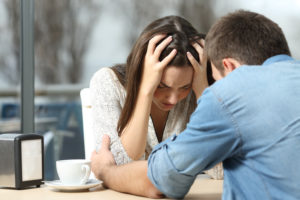 Two people sit at table in cafe, one upset, the other offering comfort and support