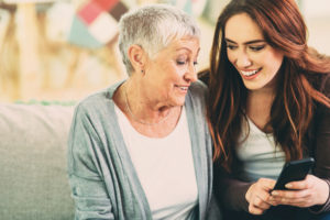 College-aged young adult with long hair sits next to short-haired grandparent. The two smile while looking at smartphone together