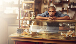 Non-white server waits behind counter of bakery/cafe for customers, resting chin on hands