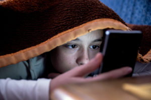 A girl is hiding under the covers as she checks her smartphone.