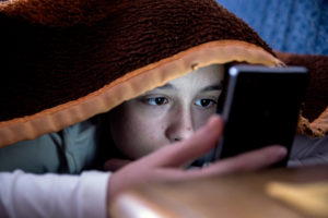 A girl is hiding under the covers as she checks her smartphone.