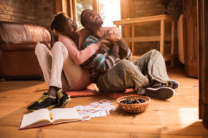 Laughing couple embraces at home, cards ant notebook and snacks next to them on floor